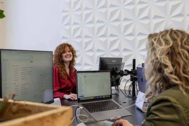Twee vrouwen zitten tegenover elkaar aan een bureau in een kantoorruimte. De vrouw links met krullend rood haar glimlacht. De vrouw op de voorgrond werkt op een laptop. Er zijn verschillende computerschermen te zien op het bureau. De ruimte heeft een moderne, geometrische wanddecoratie.